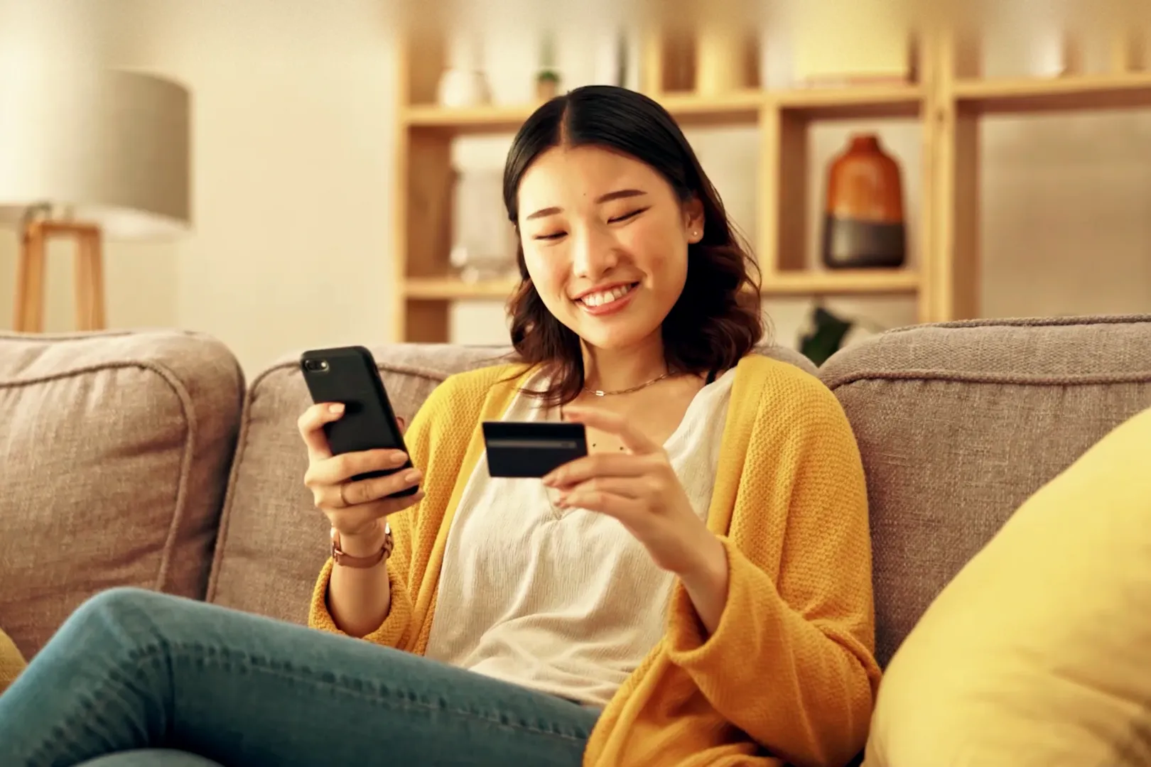 A young woman smiling at her phone, holding a card, on a sofa at home.